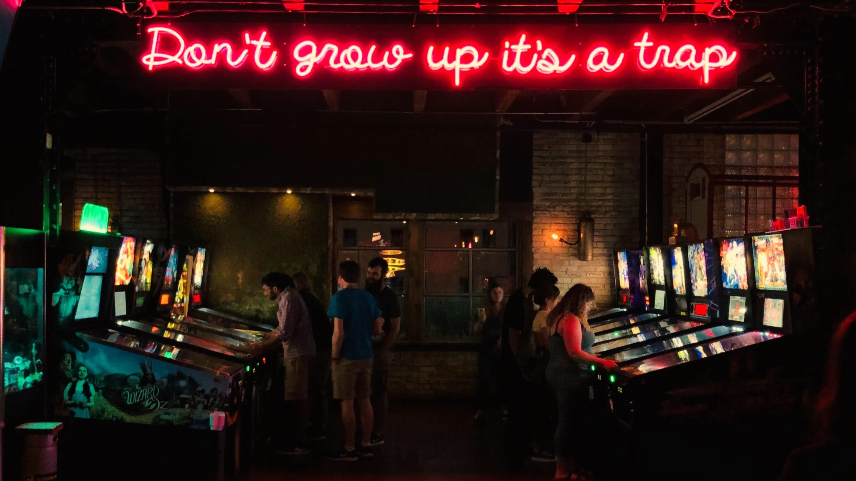 A game hall with a lot of arcade machines, people talking and a glowing neon text saying 'Don´t grow up its a trap'